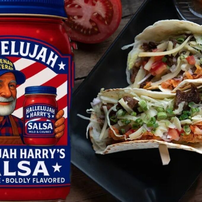 Two loaded tacos next to a jar of salsa and a sliced tomato.